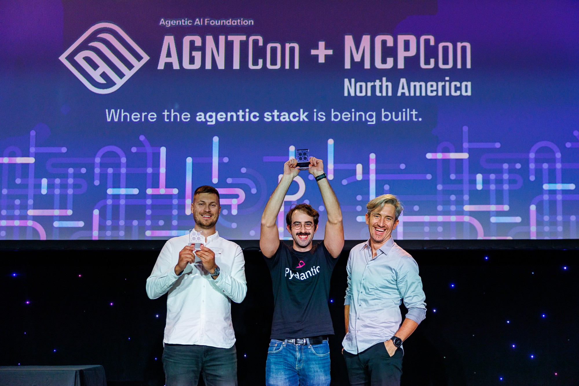 Marcelo Trylesinski holding the AAIF Outstanding Technical Contribution Award above his head on the AGNTCon + MCPCon North America stage, between Antoni Kozelski and Bartosz Gonczarek from Vstorm